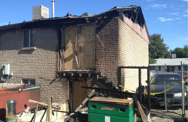 Exterior of a two-story brick building with severe fire damage, including charred siding, missing staircase, and construction debris.