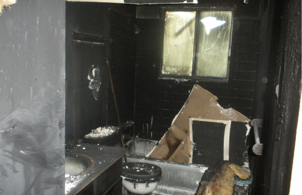 Severely fire-damaged bathroom with charred walls, broken window, melted fixtures, and debris scattered throughout. The space is blackened with soot and heavily destroyed.