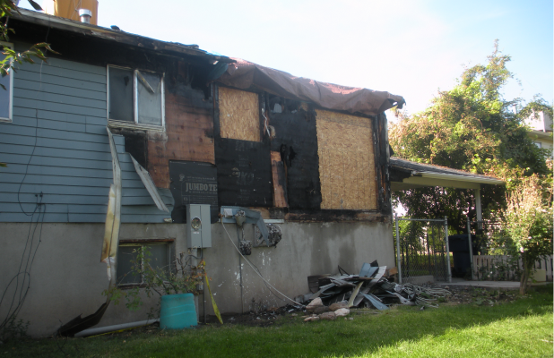 Exterior view of a residential home showing severe fire damage. The siding is burned away in sections, windows are broken, and the wall is partially boarded up with plywood. Debris is scattered on the ground near the house.