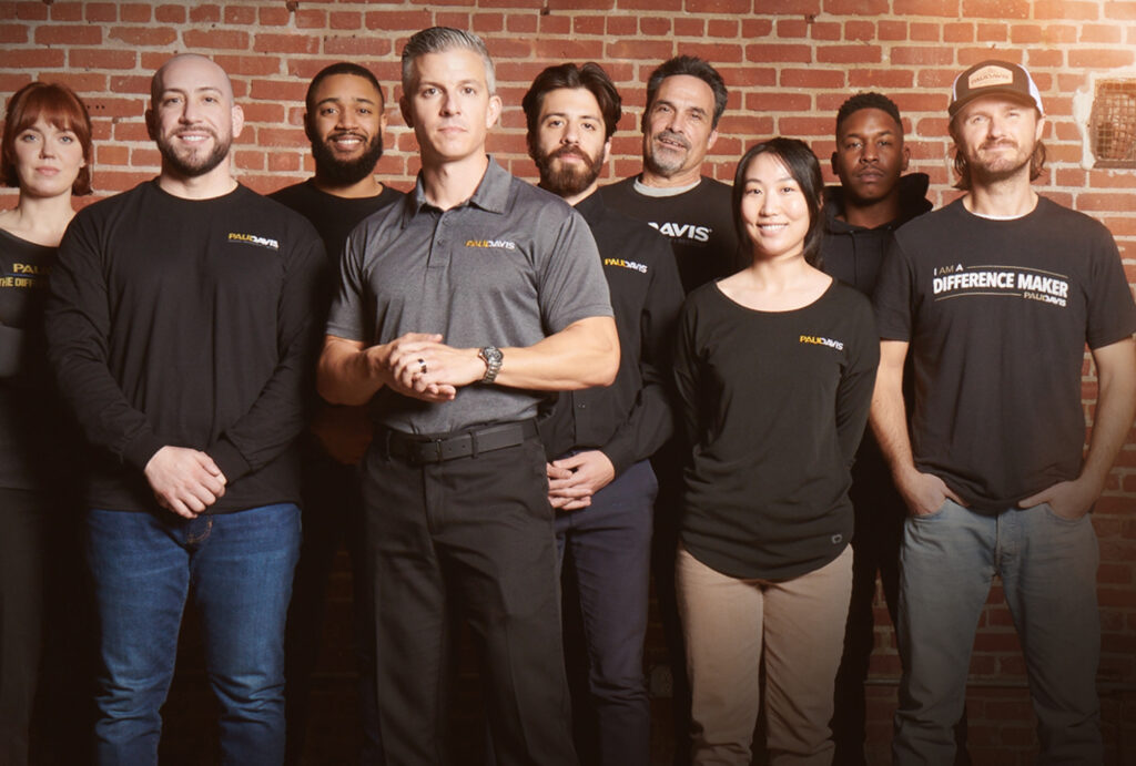 Group portrait of Paul Davis team members standing in front of a brick wall, wearing branded shirts and smiling confidently.