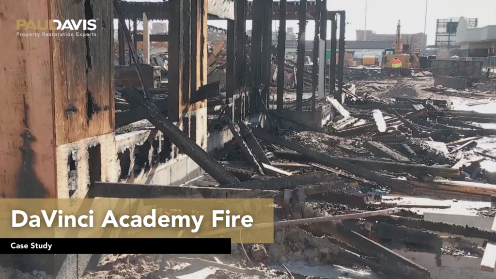 Severe fire damage at DaVinci Academy with charred wooden beams, collapsed structures, and debris scattered across the construction site.