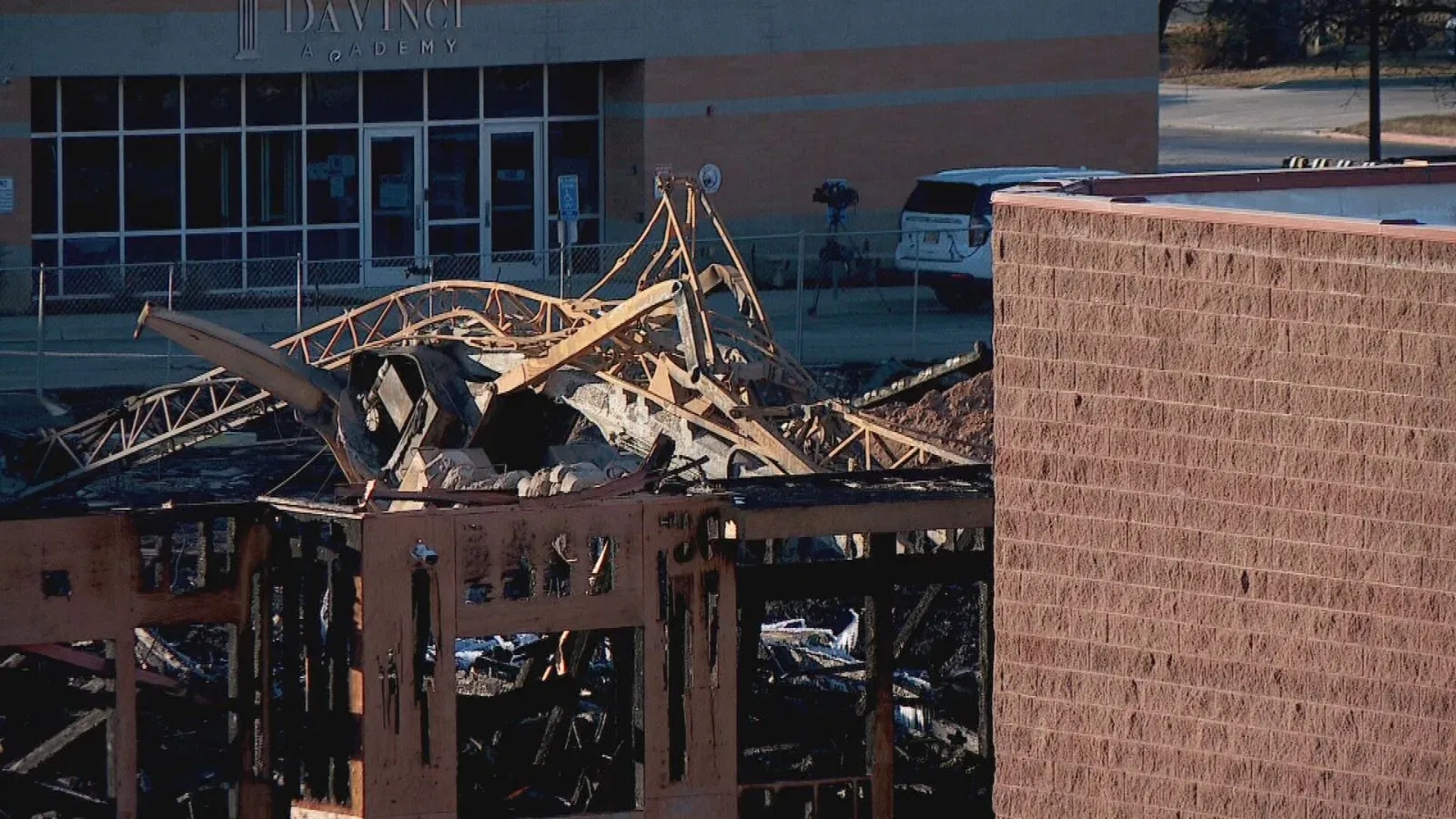 Charred remains of a commercial building with collapsed wooden roof trusses, located near an intact brick structure and a storefront.